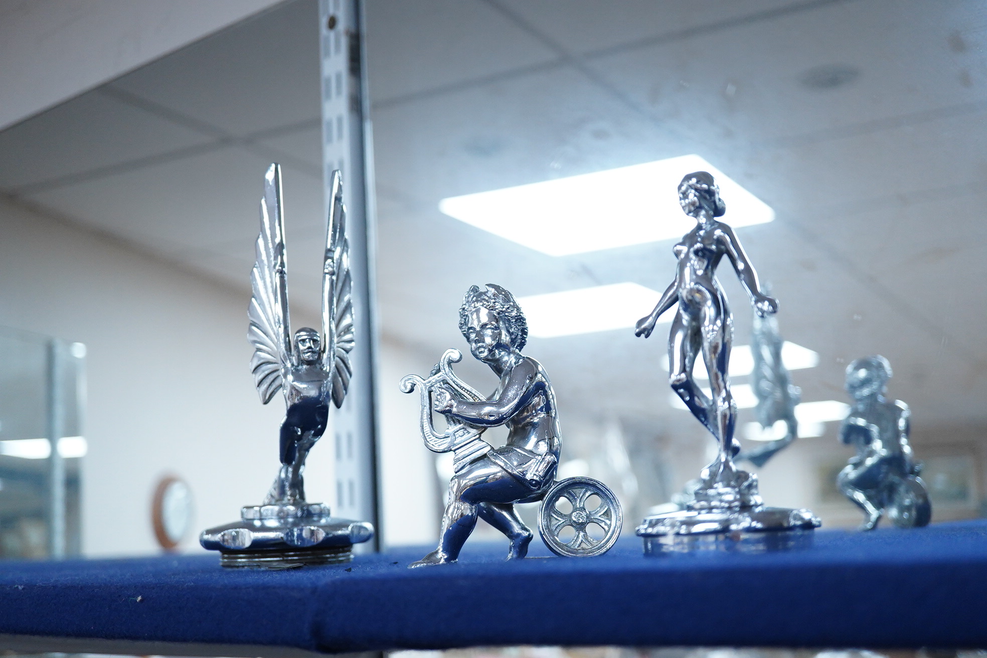 Six car mascots including five chrome and brass examples; a female figure, a cherub with harp, an Art Deco winged figure, etc. and a cold painted spelter figure of a skier
